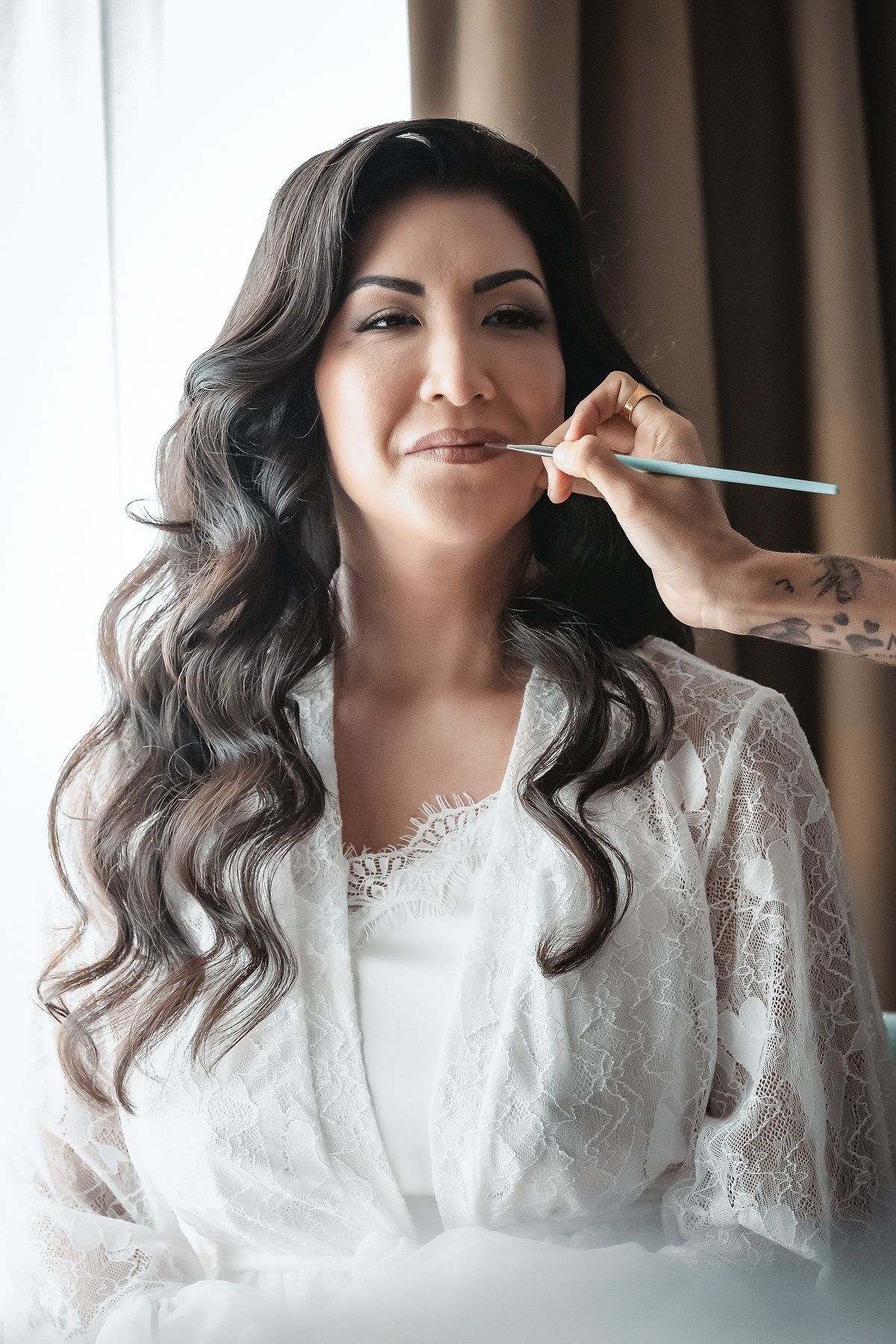 Bride in white lace robe getting makeup applied by artist with tattooed hand near bright window before wedding ceremony.