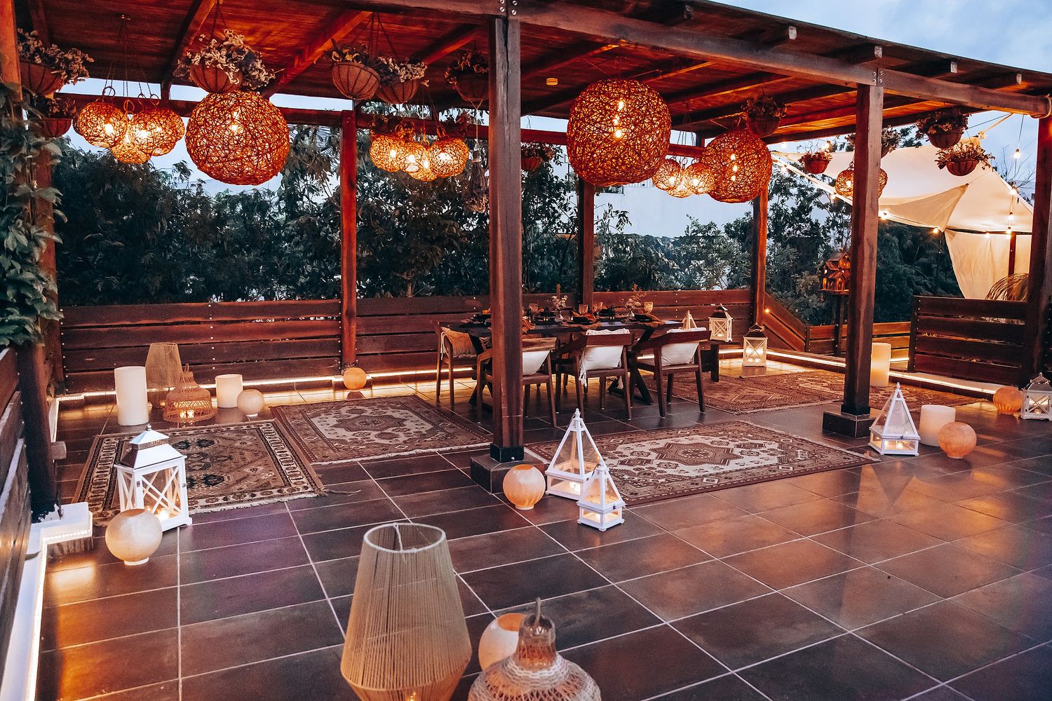 Romantic boho outdoor terrace dining setup with wicker pendant lights, lanterns, and patterned rugs at dusk.