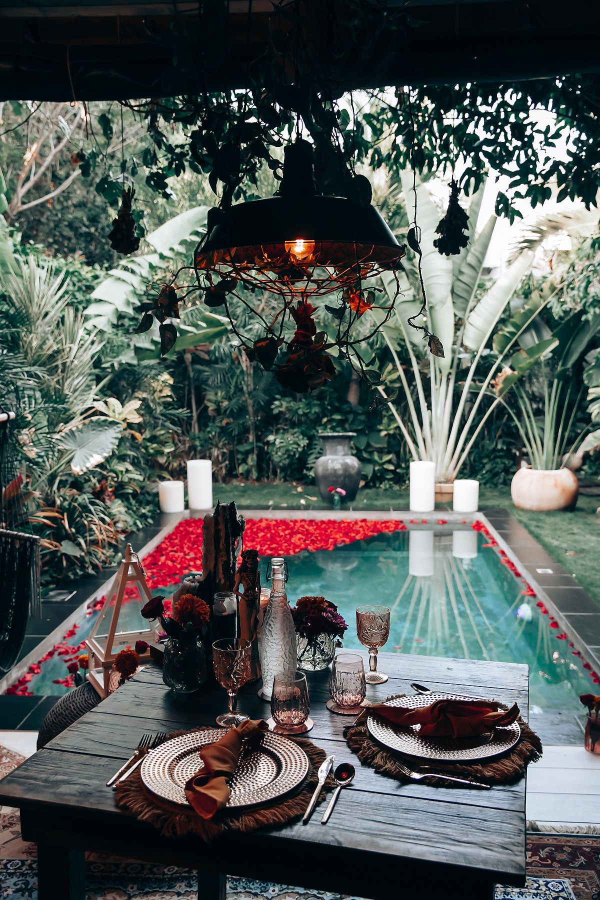 Romantic outdoor dining table set for two beside a flower-petal pool, surrounded by lush tropical greenery and candlelight.