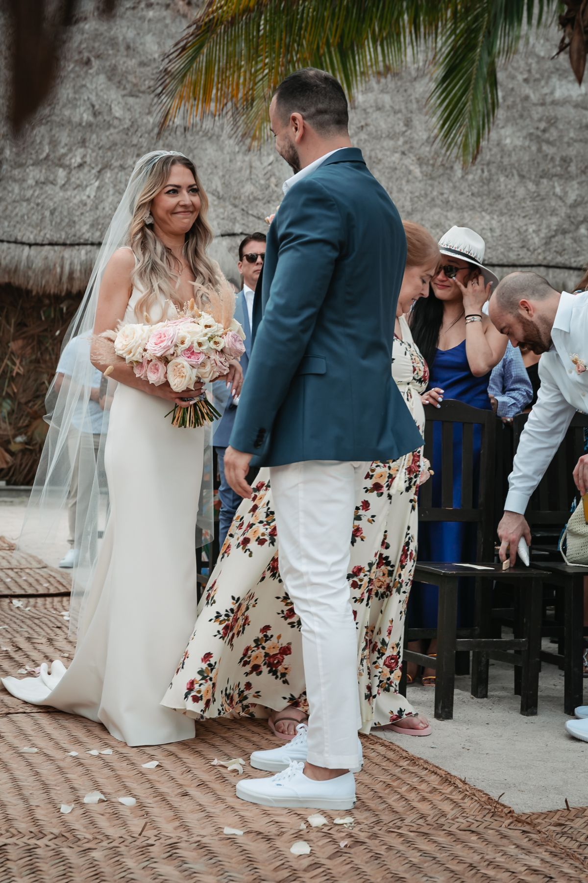 Bride in white gown holding pink bouquet smiles at groom during outdoor beach wedding ceremony with guests.