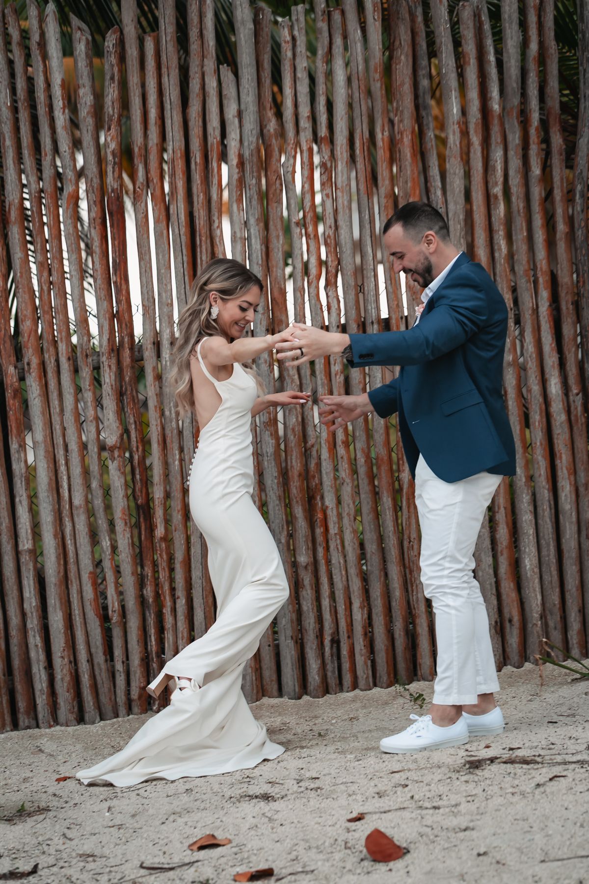 Bride and groom dancing playfully on sandy beach in front of rustic wooden fence.