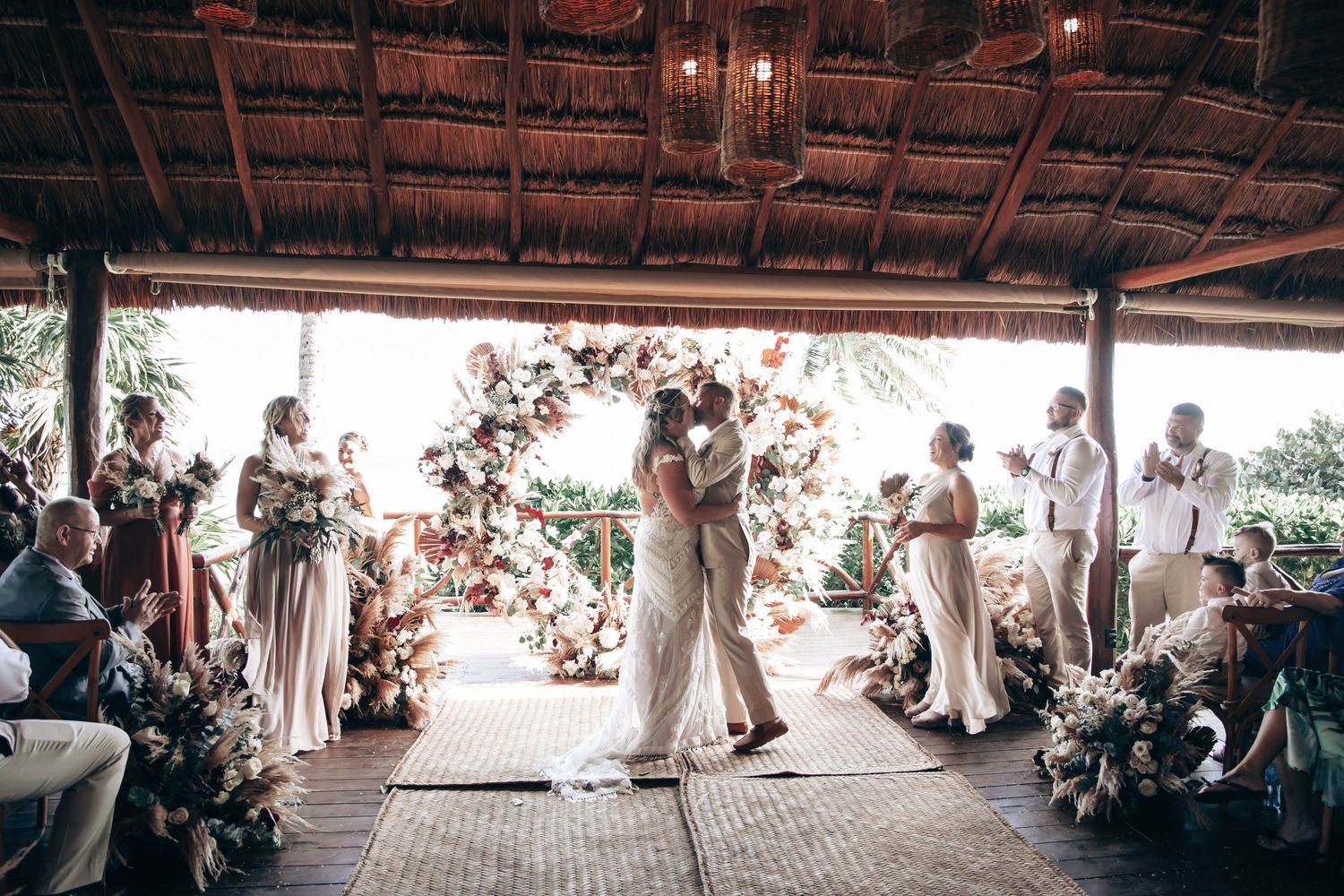 Bride and groom sharing first kiss under floral arch at tropical wedding ceremony.