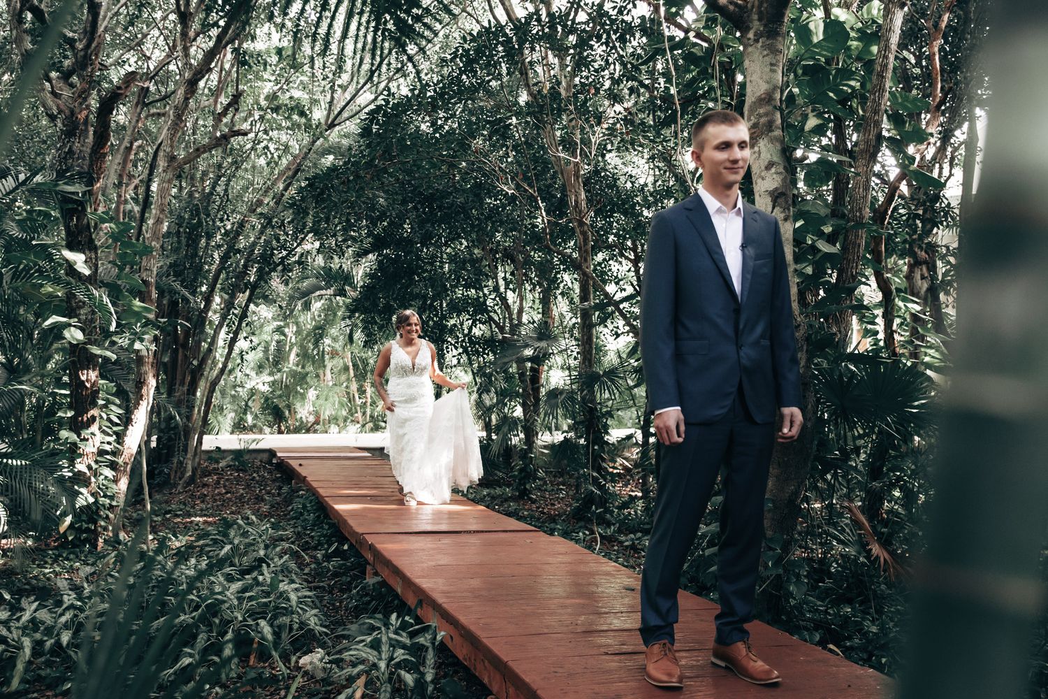 Groom in navy suit standing on wooden boardwalk as bride in white dress approaches through tropical forest setting.