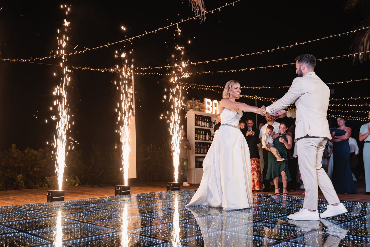 Bride and groom dance on mirrored floor with sparkling pyrotechnic fountains at reception.
