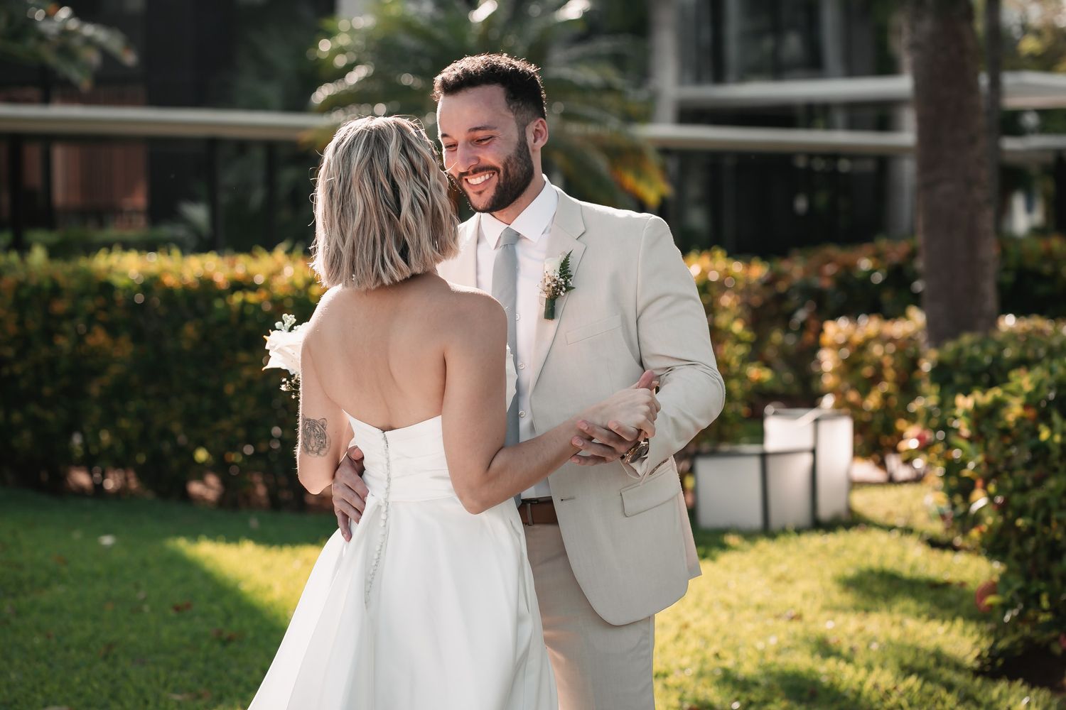 Groom in beige suit smiling warmly at bride during outdoor first look moment.