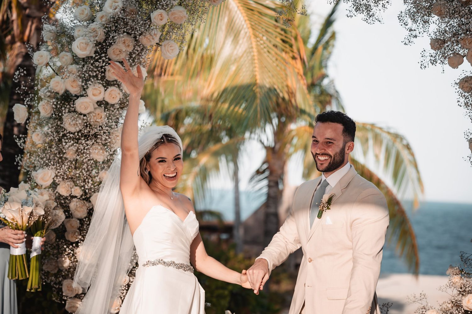 Bride and groom celebrate at outdoor wedding ceremony with floral arch and palm trees.