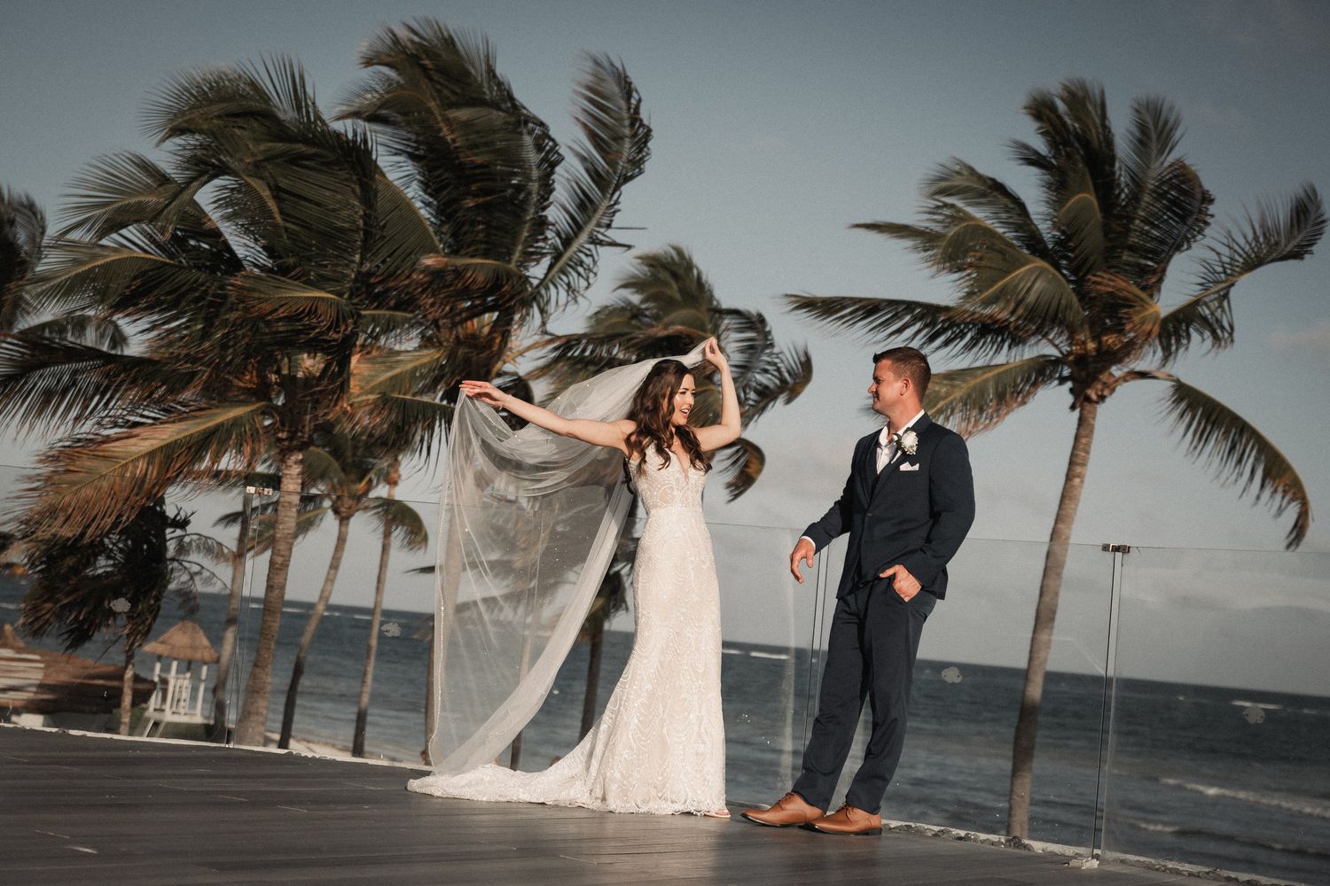 Bride and groom dance on beachside deck with palm trees swaying in the wind.