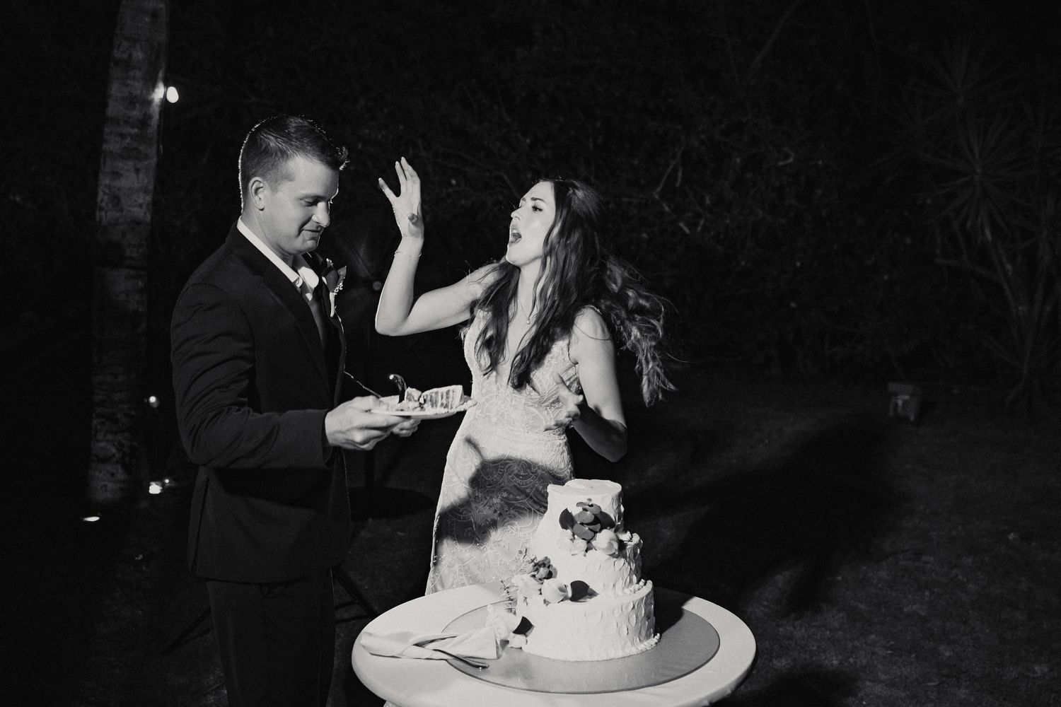 Bride and groom laugh while cutting their wedding cake at an outdoor nighttime reception in black and white photo.