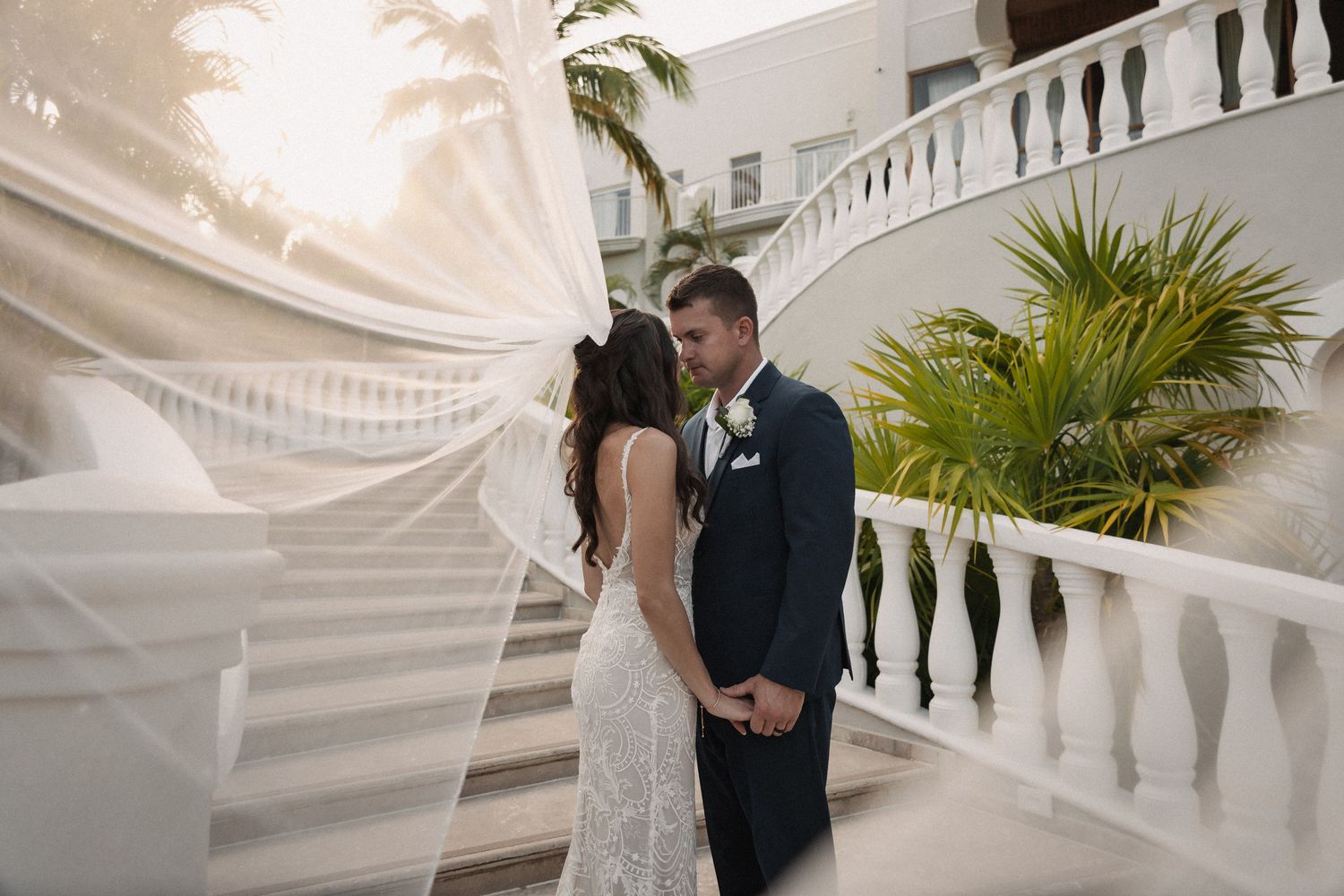 Bride and groom share a romantic moment on white staircase with veil flowing in the breeze at tropical wedding venue.
