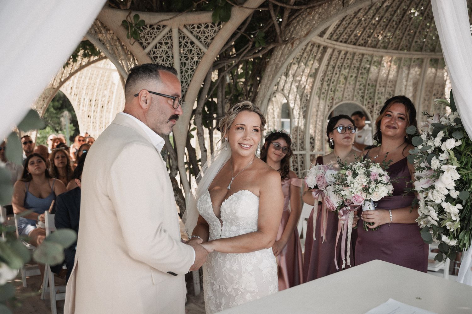 Smiling bride in strapless gown exchanges vows with groom at outdoor wedding ceremony with bridesmaids.