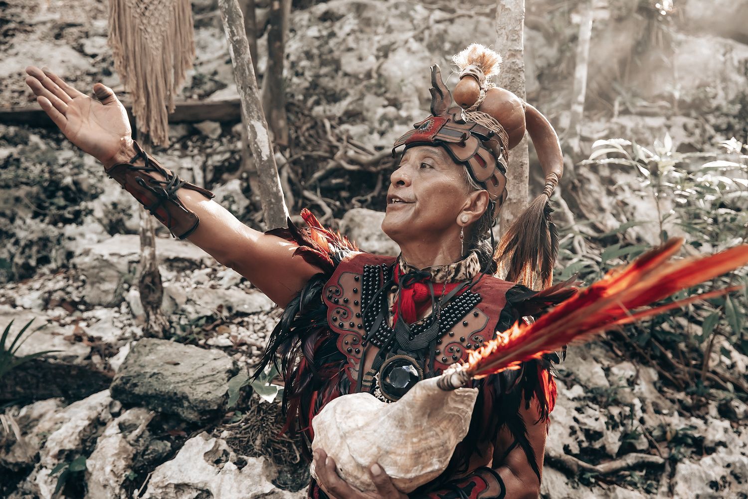 A woman wearing a colorful, feathered headdress and traditional clothing stands in a rugged, natural setting.