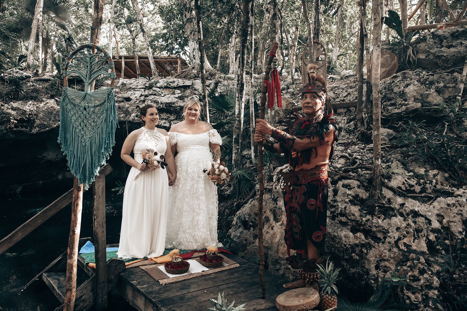 Two women in white dresses stand on a wooden bridge in a forest, with a person in a red costume nearby.