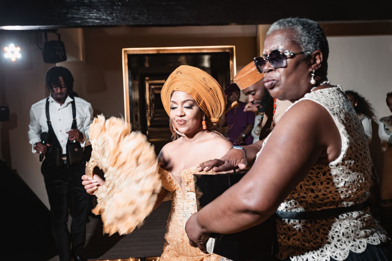 Bride in stunning orange Nigerian traditional attire dances with a feather fan at her wedding celebration.