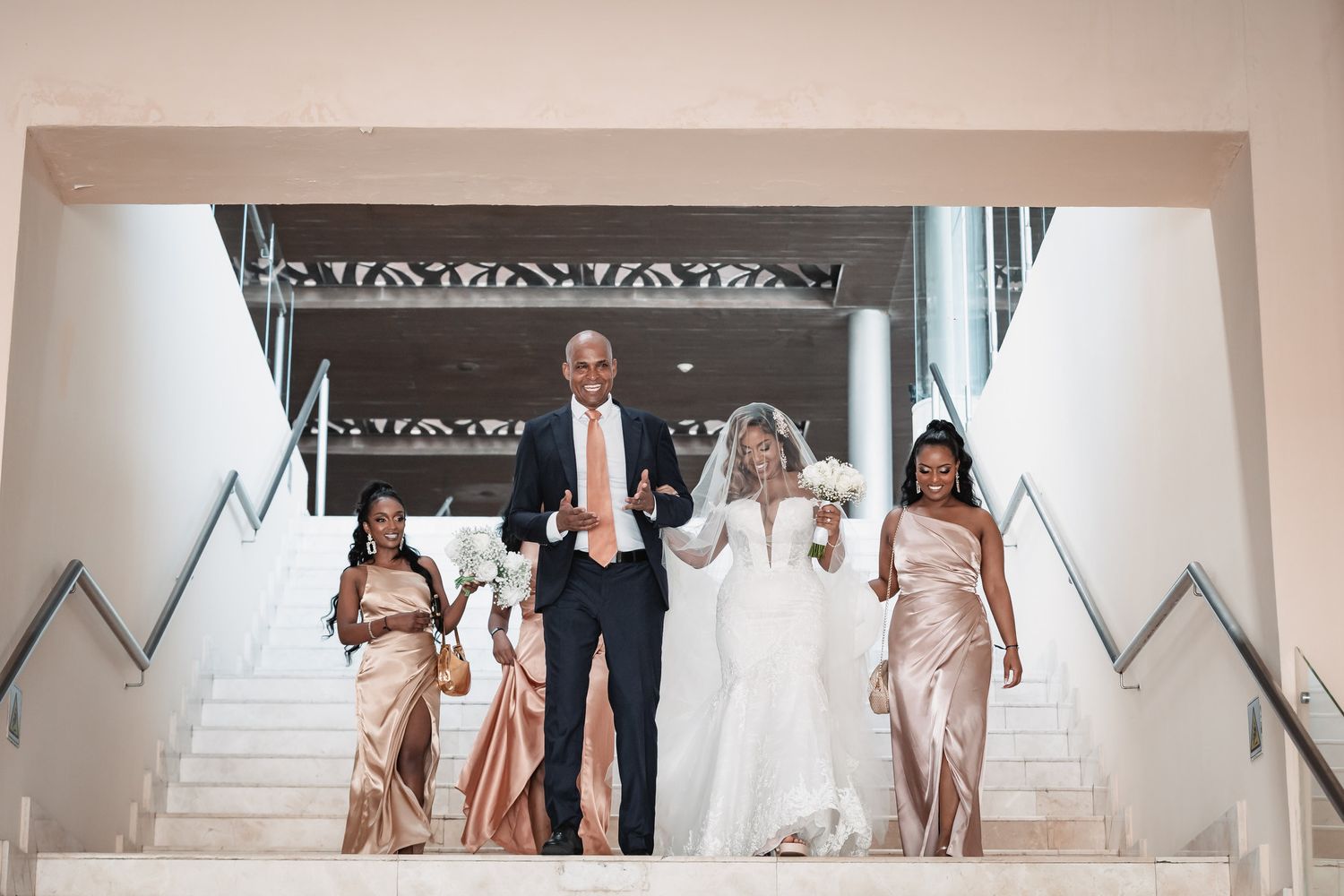 Bride in white lace gown descending grand staircase with family and bridesmaids in gold dresses.