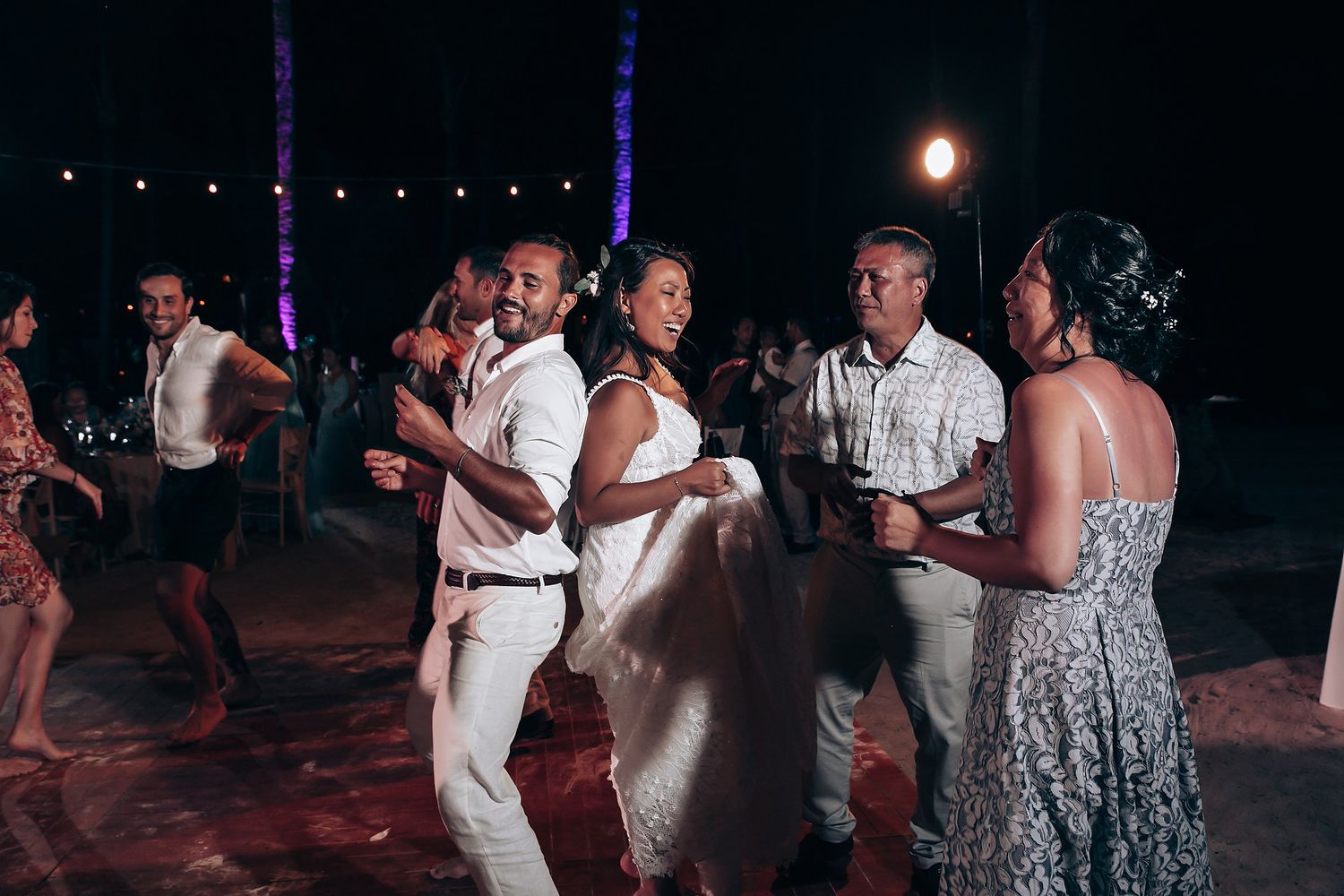 Wedding party dances under blue lighting and string lights during an evening celebration.
