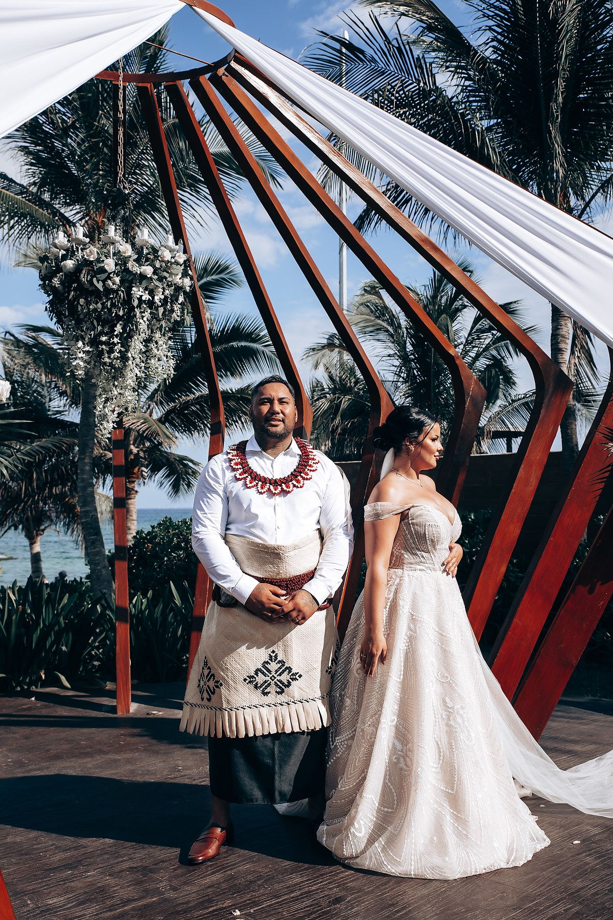Wedding couple posing under a wooden pergola structure with white fabric draping and palm trees.