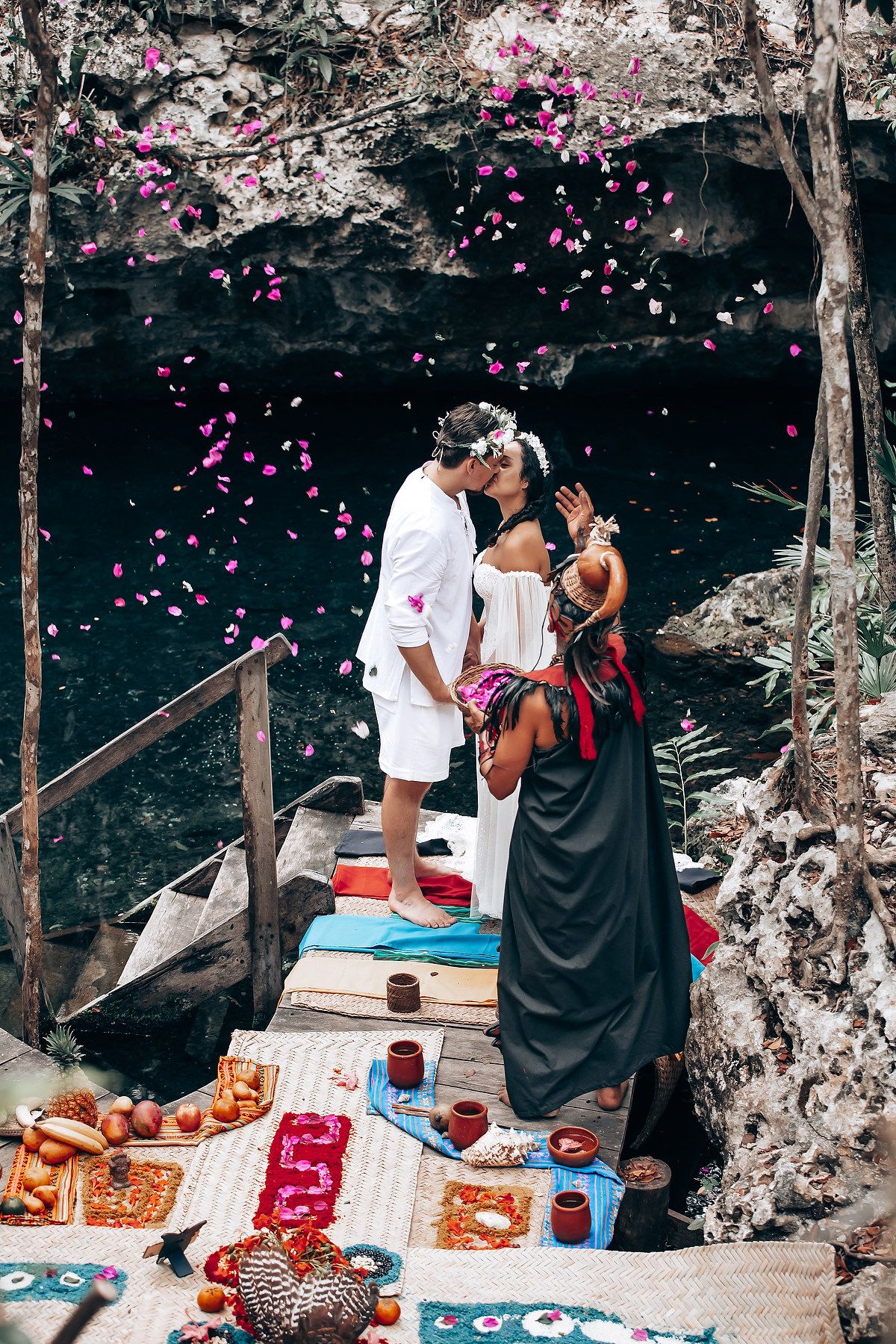 Romantic picnic setup with pink flower petals falling near a cave entrance, featuring blankets and food spread on wooden deck.