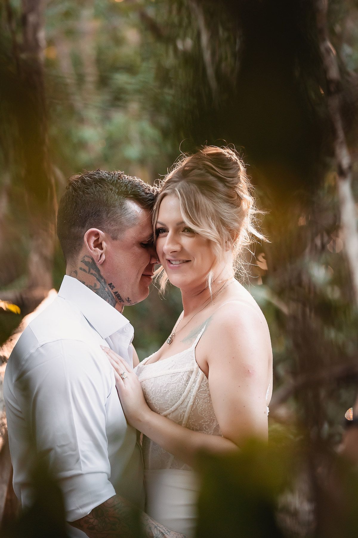 Couple embracing intimately in a lush forest setting, bride in white dress smiling, groom with neck tattoos in white shirt.