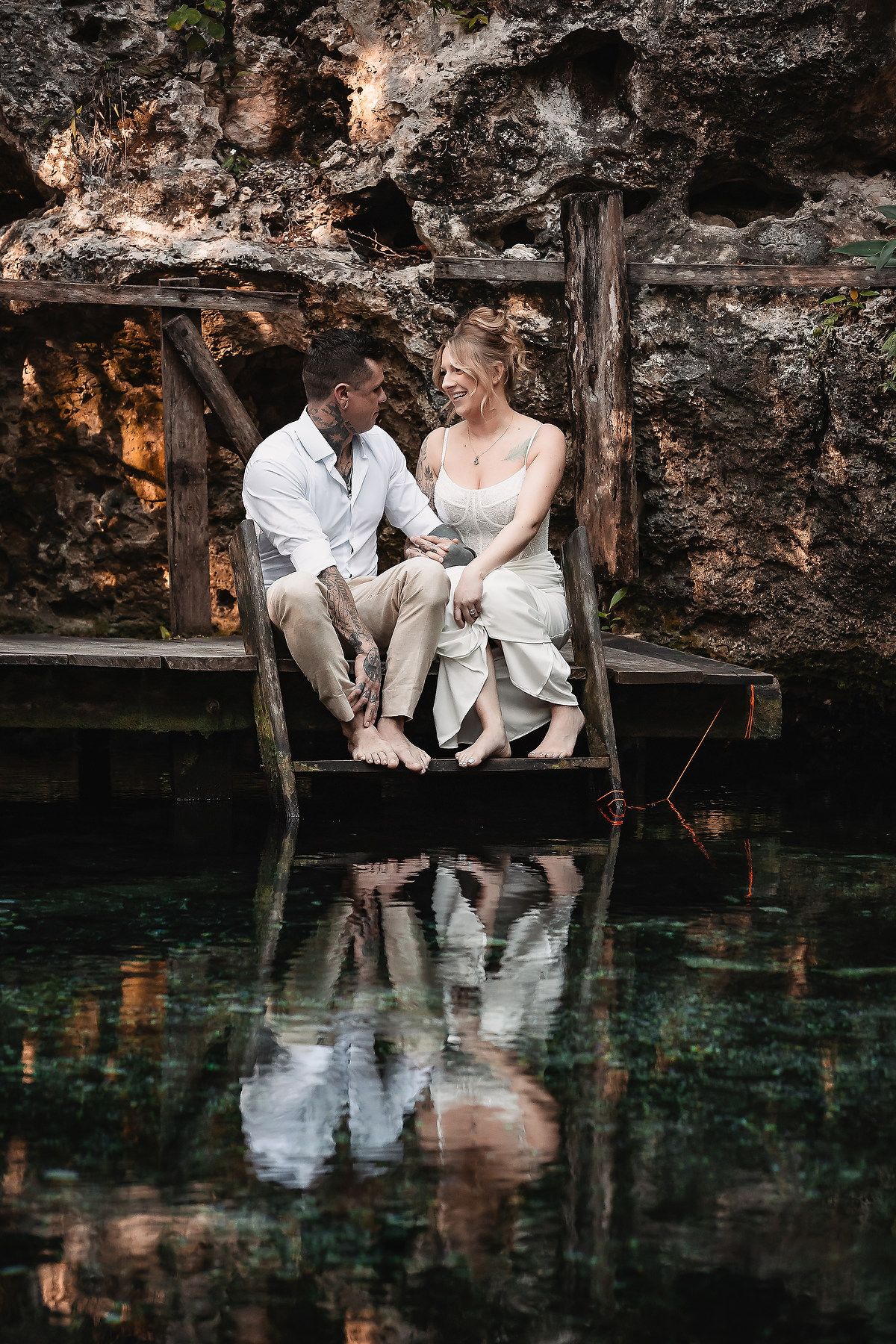 Couple in wedding attire sitting on a wooden dock over a crystal-clear cenote, sharing a romantic moment.