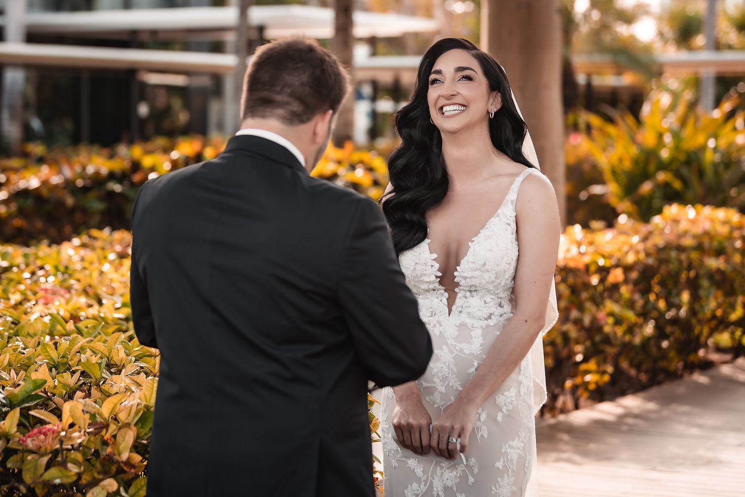Bride in lace gown smiles joyfully at groom during outdoor first look at sunny Florida wedding venue.