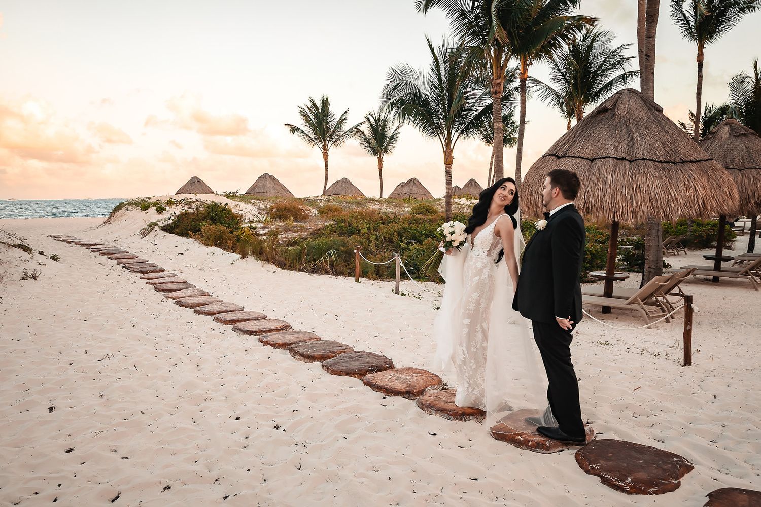 Bride and groom share a romantic sunset moment on a tropical beach with palm trees and thatched palapas in background.