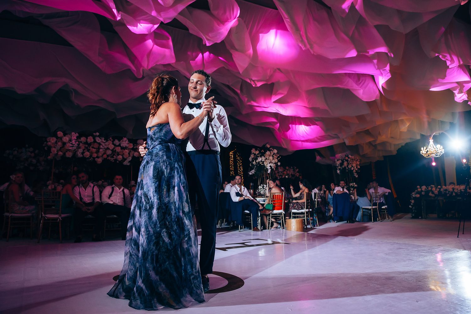 Wedding celebration with dramatic magenta lighting and white draped ceiling decorations.