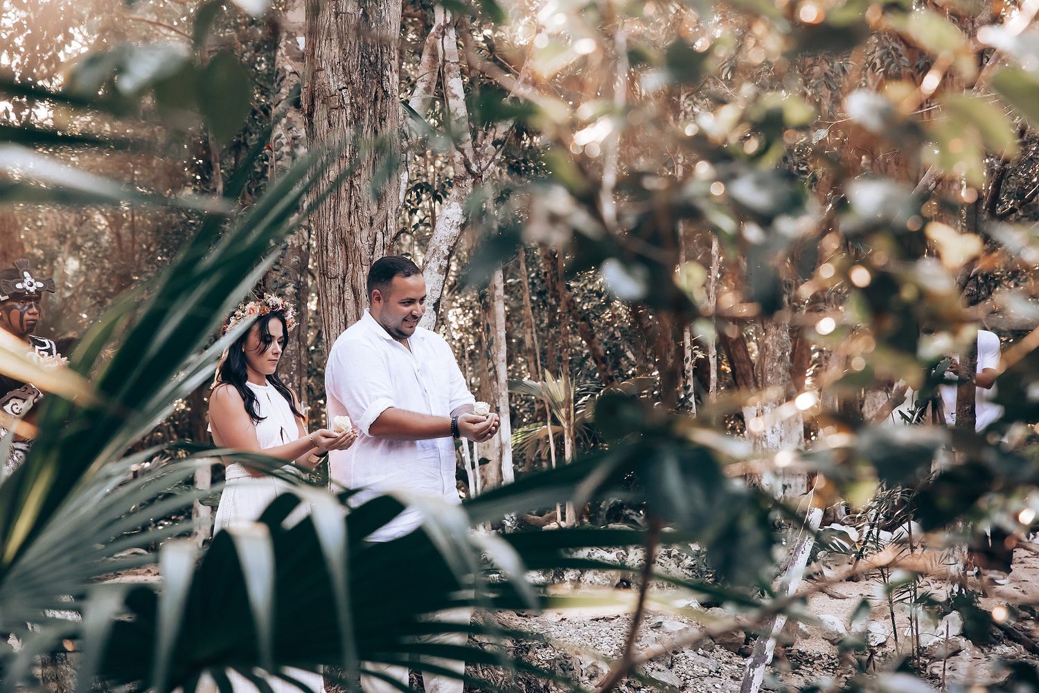 A couple in white attire stands among tropical foliage during an outdoor ceremony.