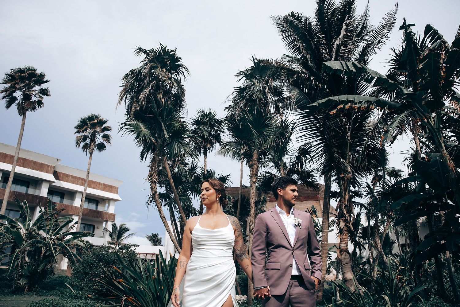 Newlyweds pose together on a palm-lined beach path in elegant attire.