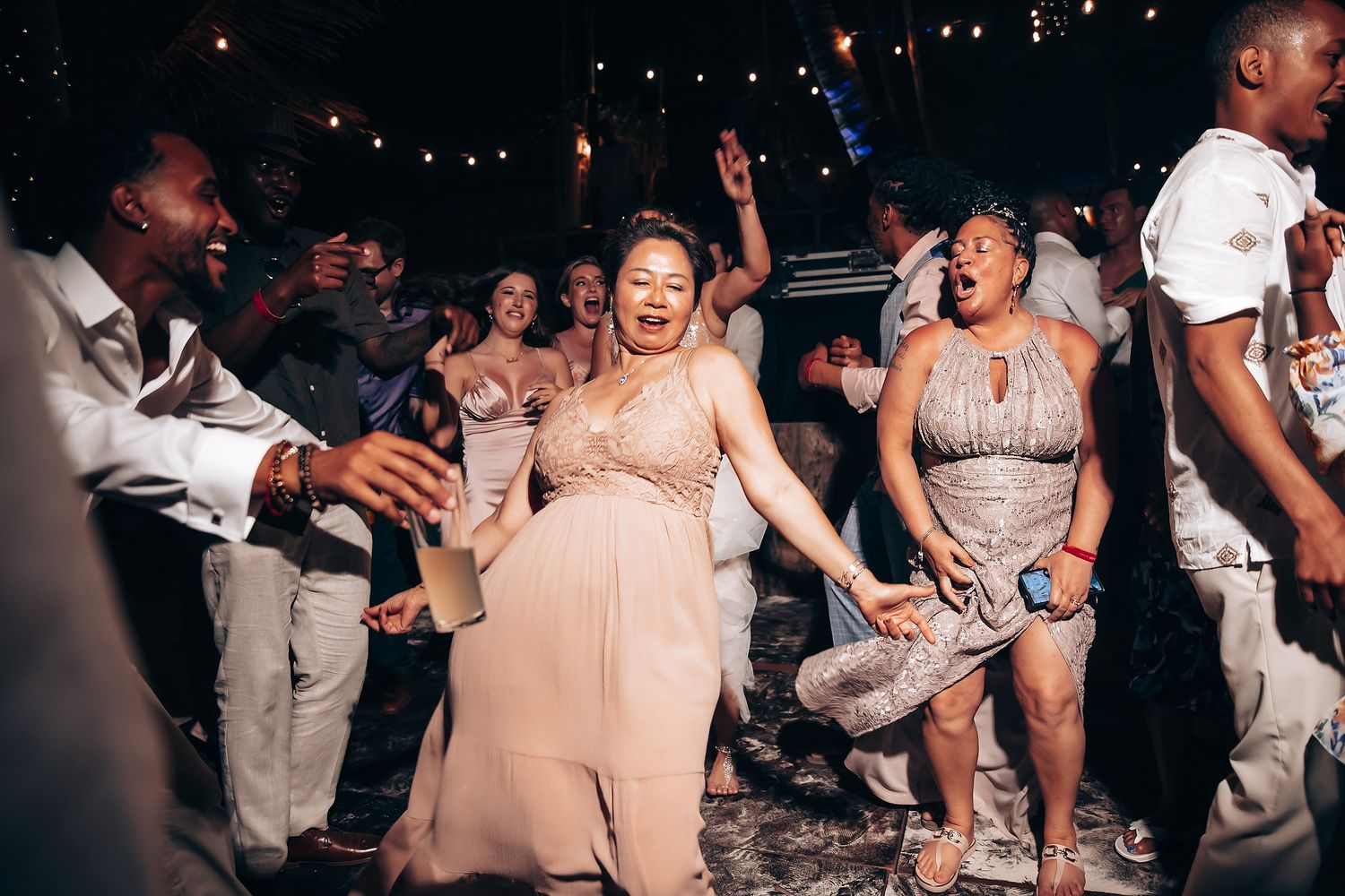 Wedding guests dancing and celebrating at a dimly lit reception with string lights overhead.
