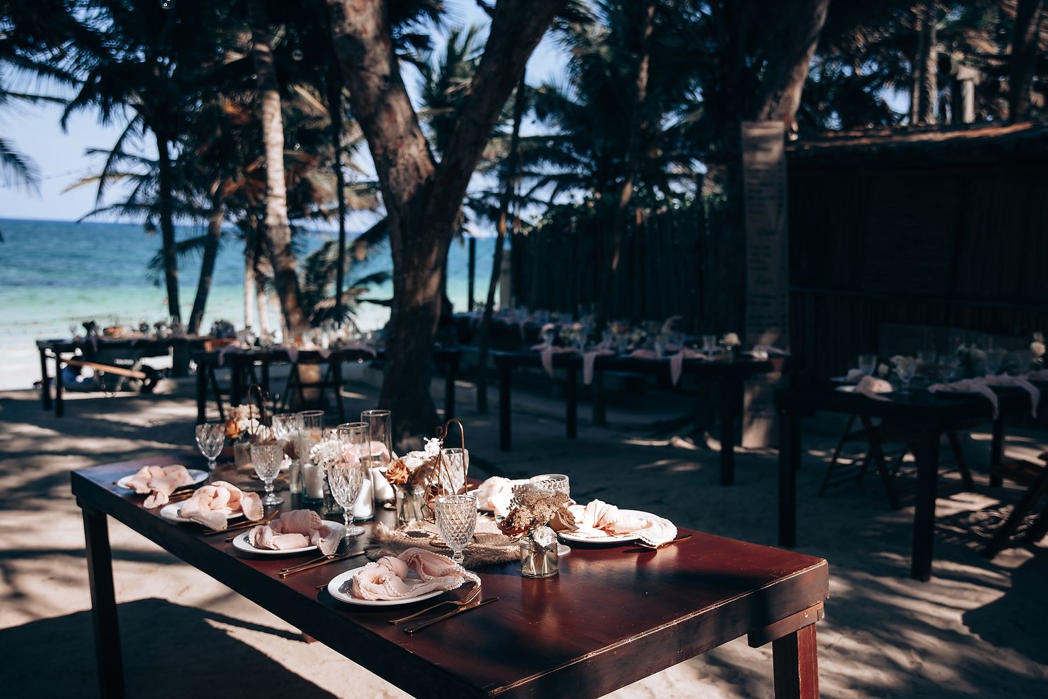 Elegant beach wedding reception setup with tables under palm trees by the ocean.