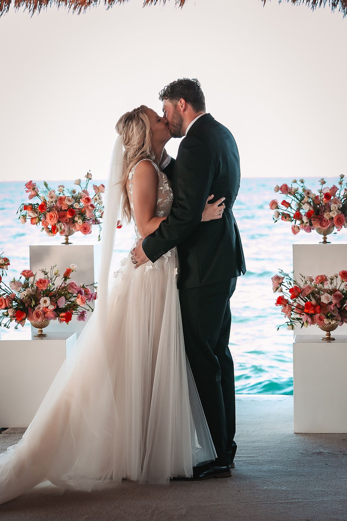 Wedding couple sharing private moment with red floral arrangements and ocean backdrop.