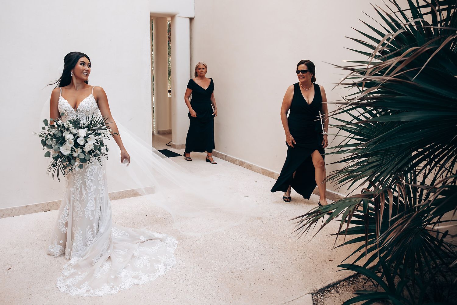 A bride in a flowing white dress walks past tropical plants with attendants in black dresses.