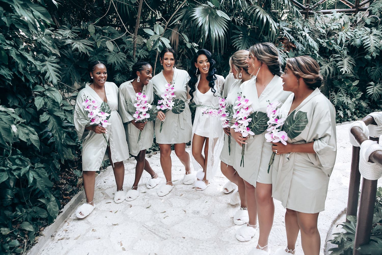 Bridesmaids in matching white robes gather together in a tropical garden setting.