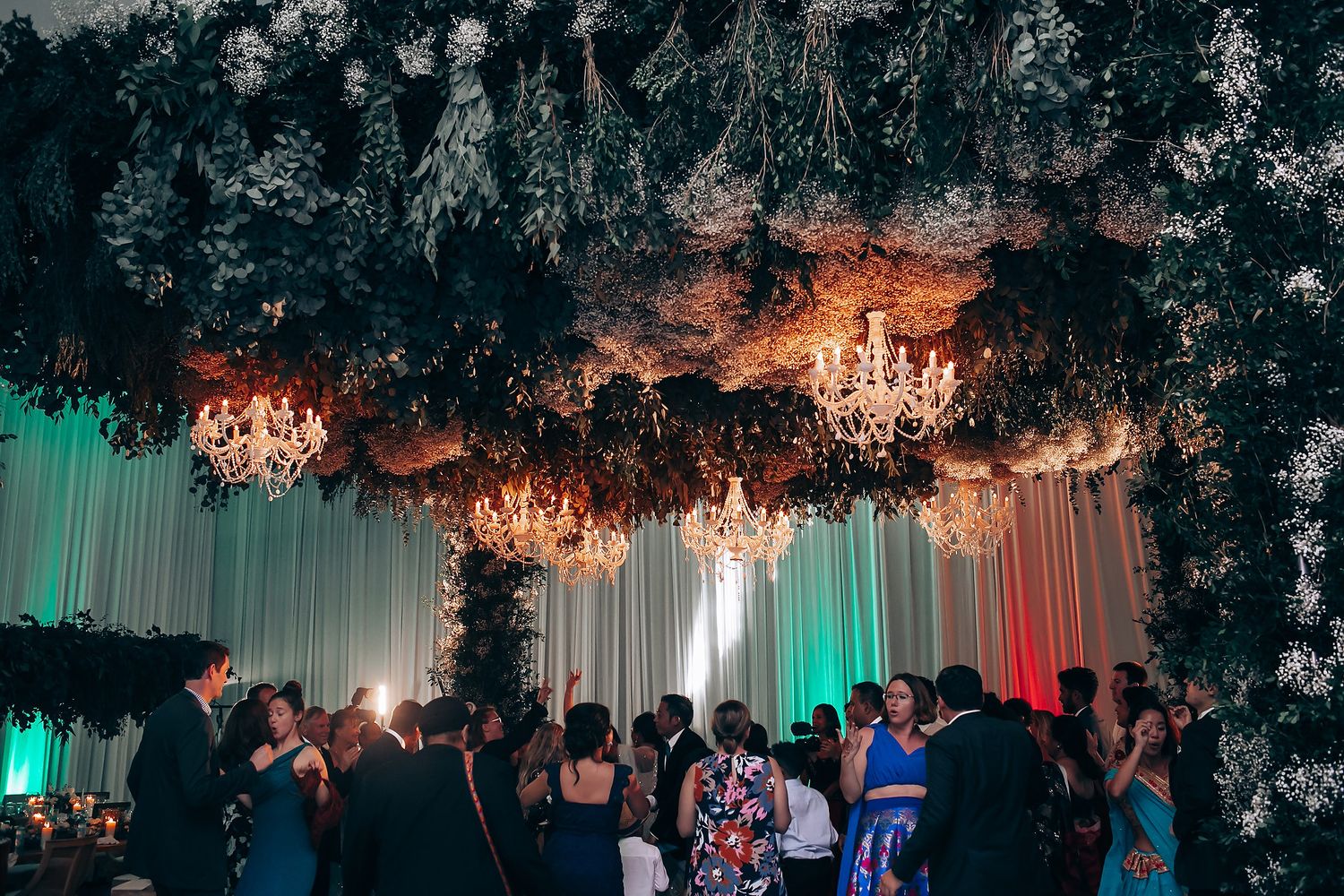 Guests gather beneath ornate chandeliers and floral decorations at an elegant indoor celebration.