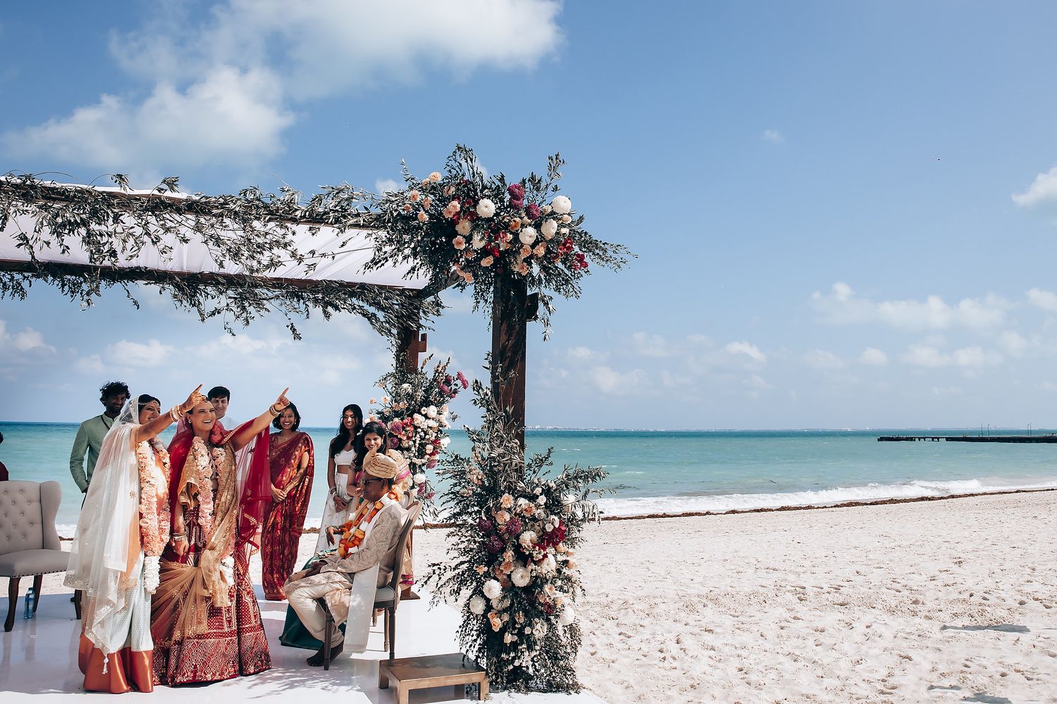 Wedding party gathered under a floral arch on a pristine beach with turquoise waters.