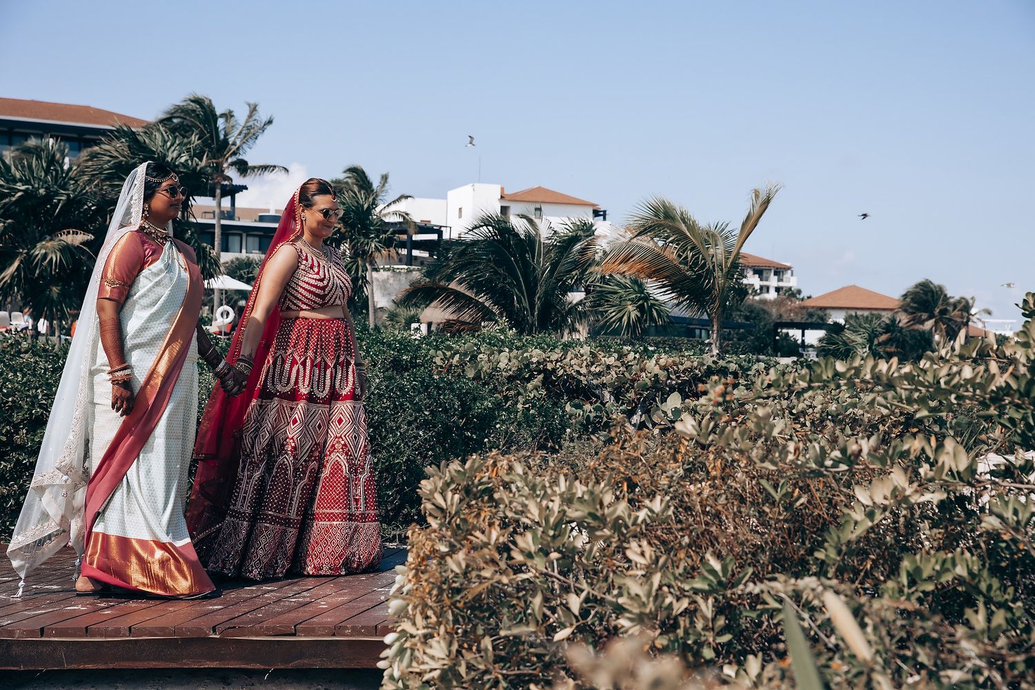 Two people in traditional Indian wedding attire walking on wooden deck with palm trees.