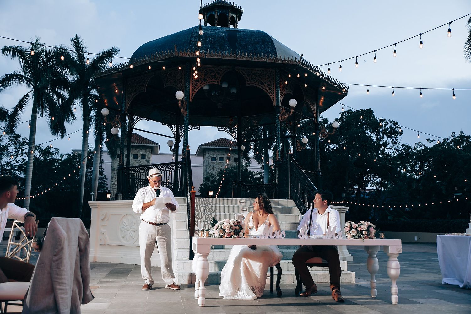Evening wedding reception under string lights at outdoor tropical venue.