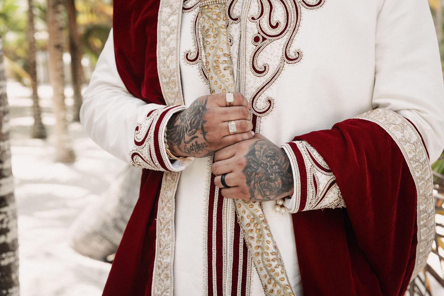 Groom in ornate white and red sherwani with gold embroidery and tattoed hands clasped at Indian wedding.