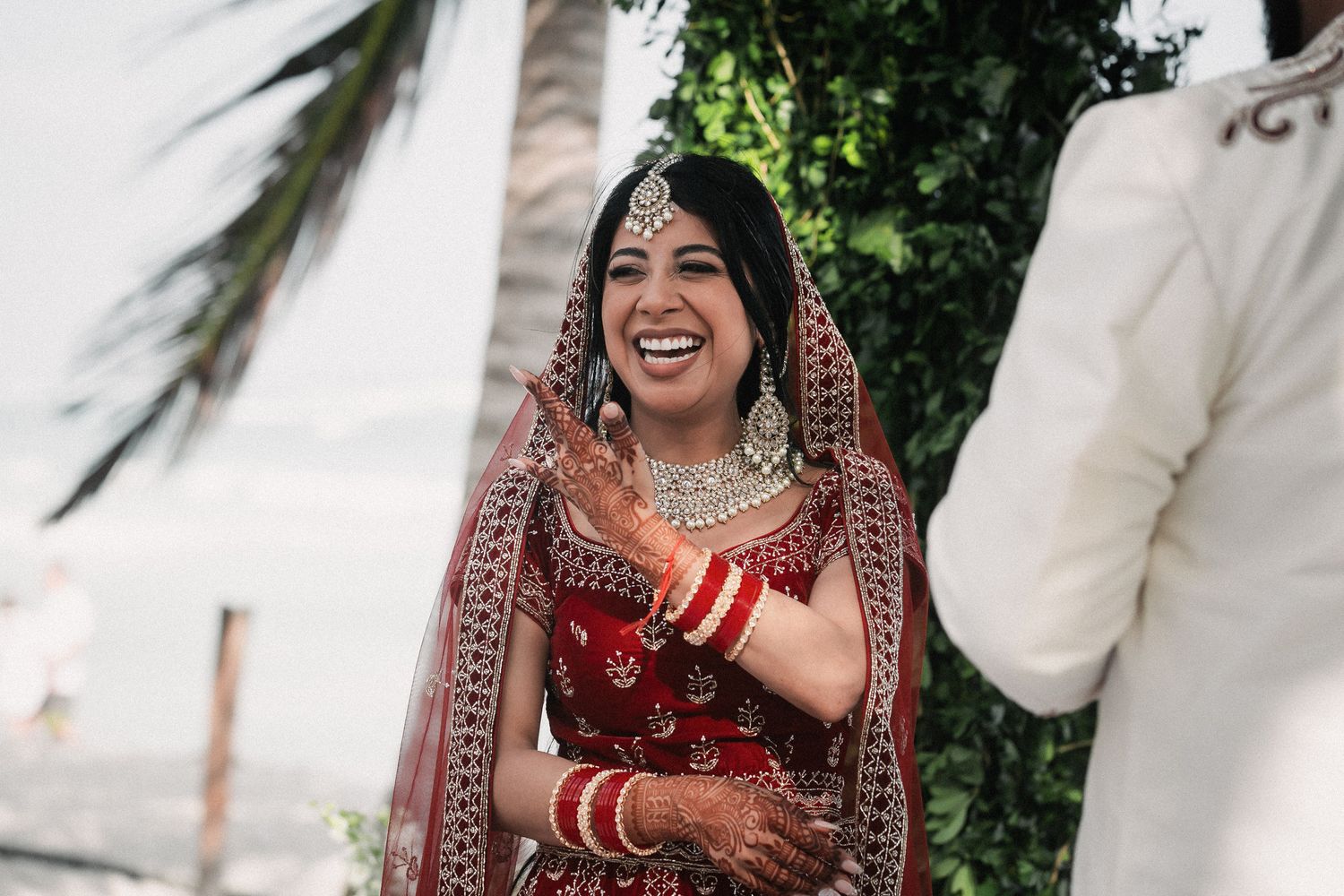 Radiant Indian bride in red bridal lehenga laughing joyfully during outdoor wedding ceremony.