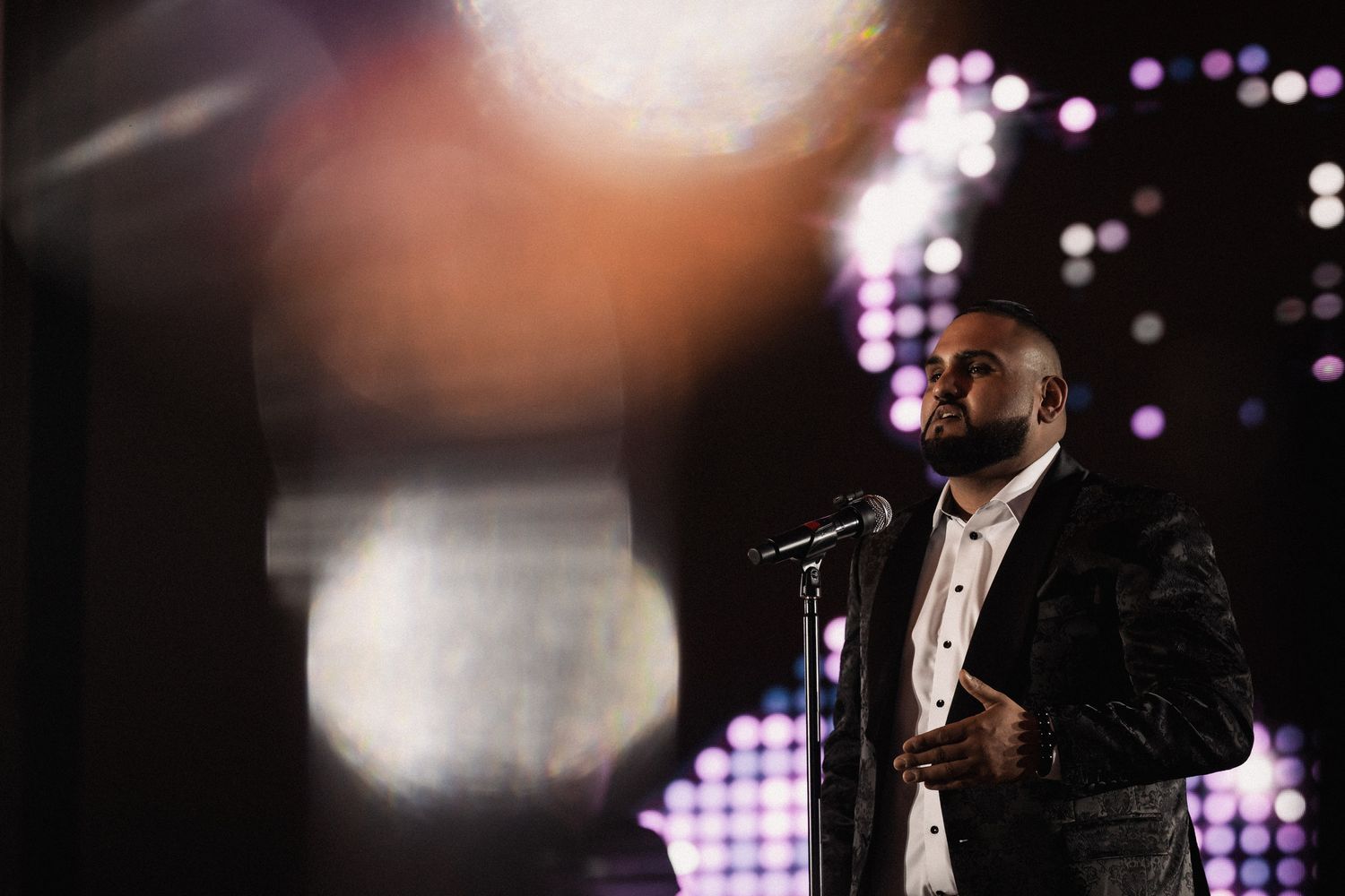 Man in sequined blazer performing at microphone on stage with purple bokeh lights in background.