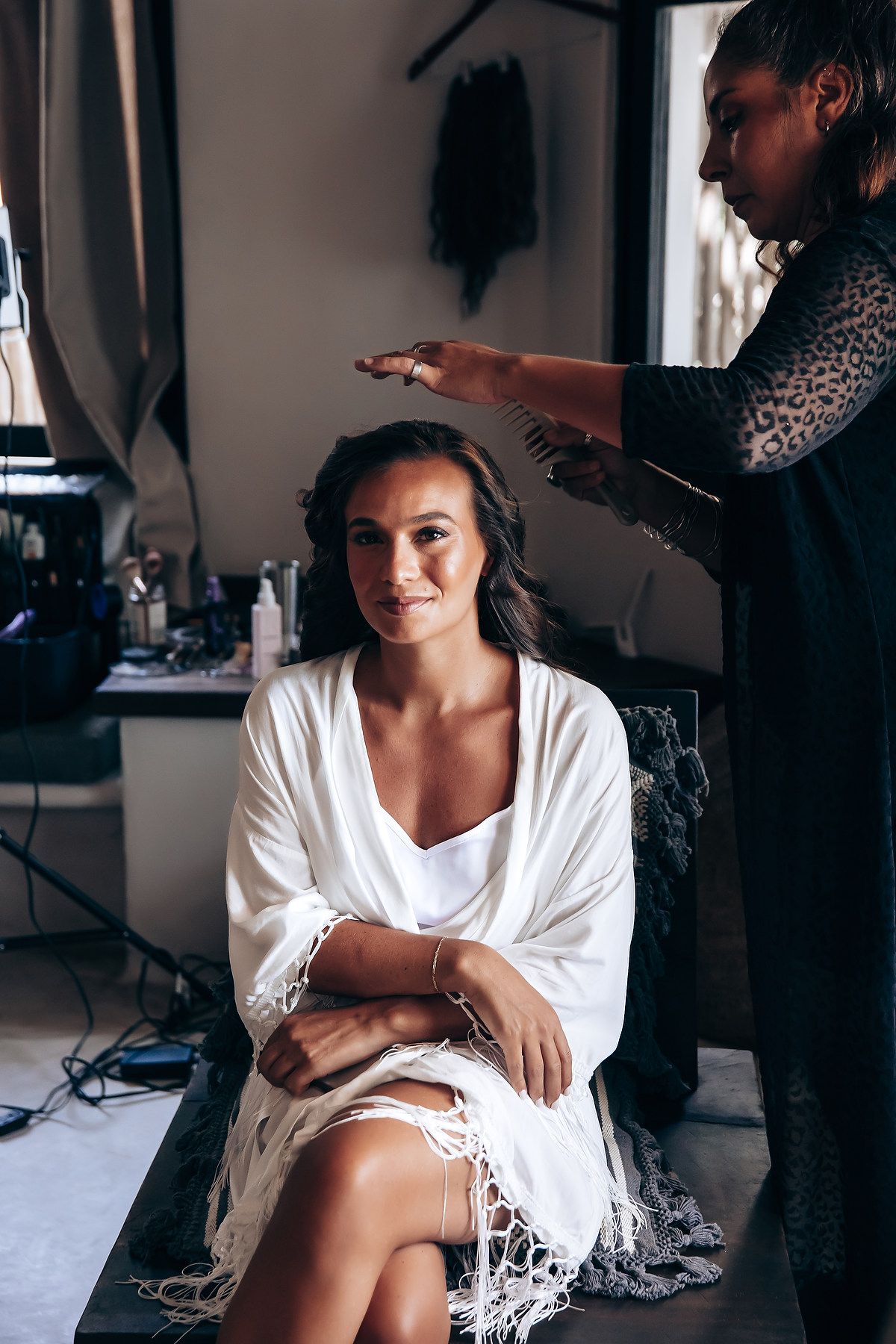 A stylist works on hair preparations in a bridal getting ready room with natural window light.