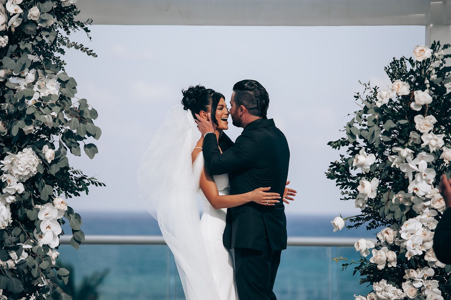 Couple sharing intimate moment during beach wedding ceremony with floral decorations.