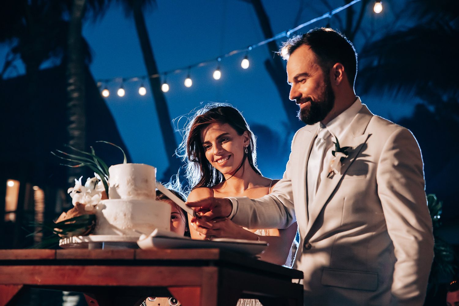 Evening wedding reception scene with string lights and cake cutting moment.