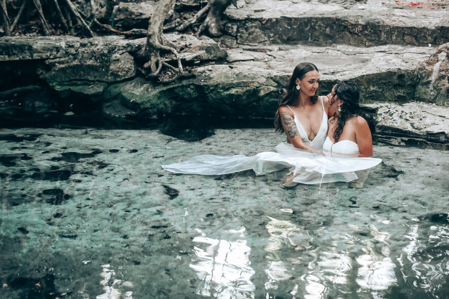 Couple in white dresses floating in clear tropical waters amid natural rock formations.