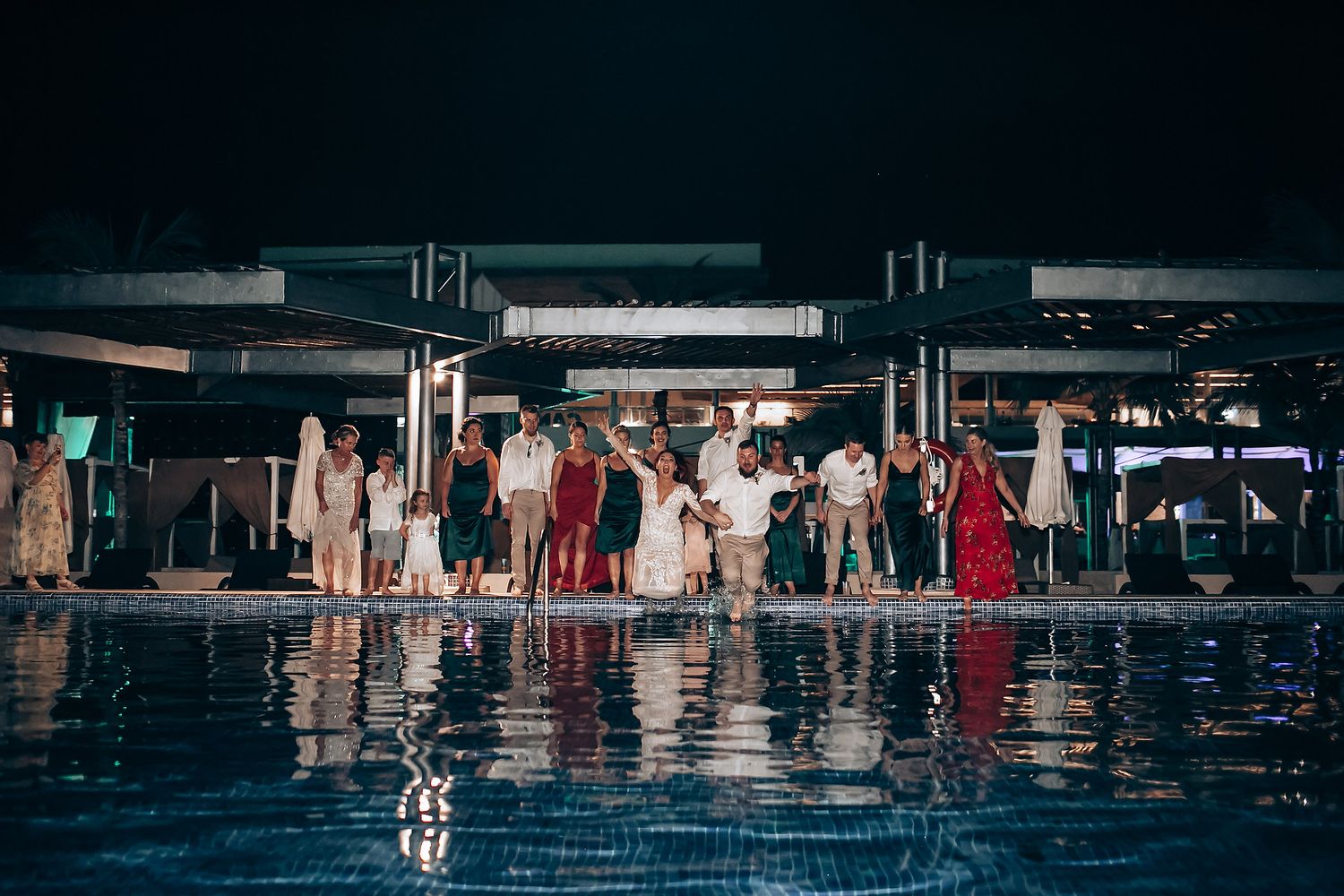 Dark reflections of wedding guests in a pool during an evening outdoor reception.