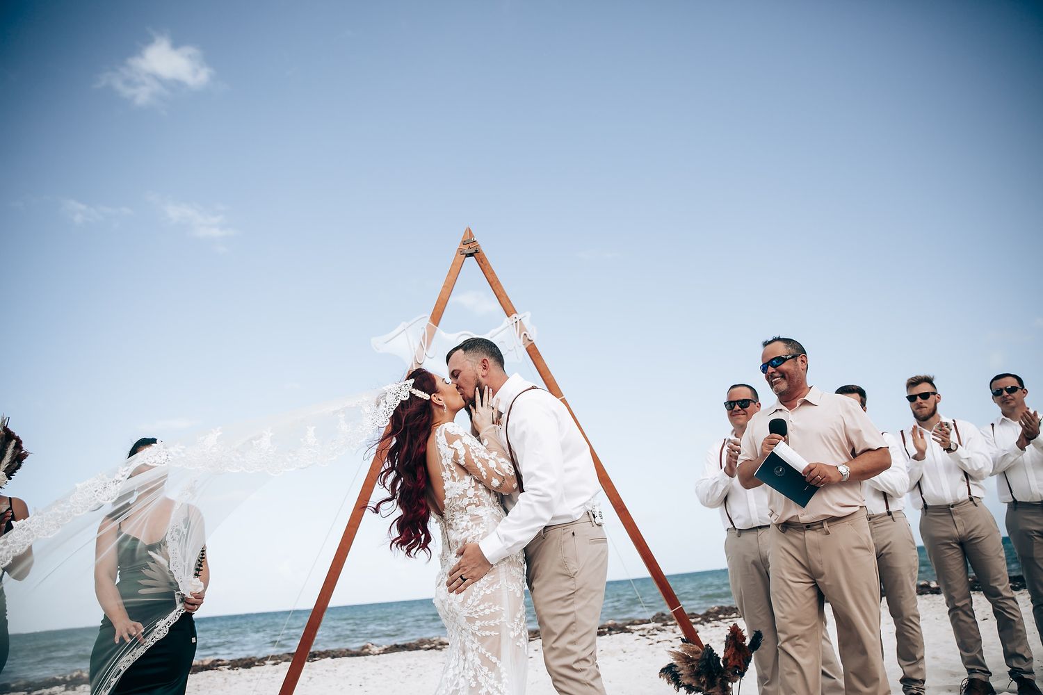 Beach wedding ceremony setup with wooden triangle arch against blue ocean backdrop.