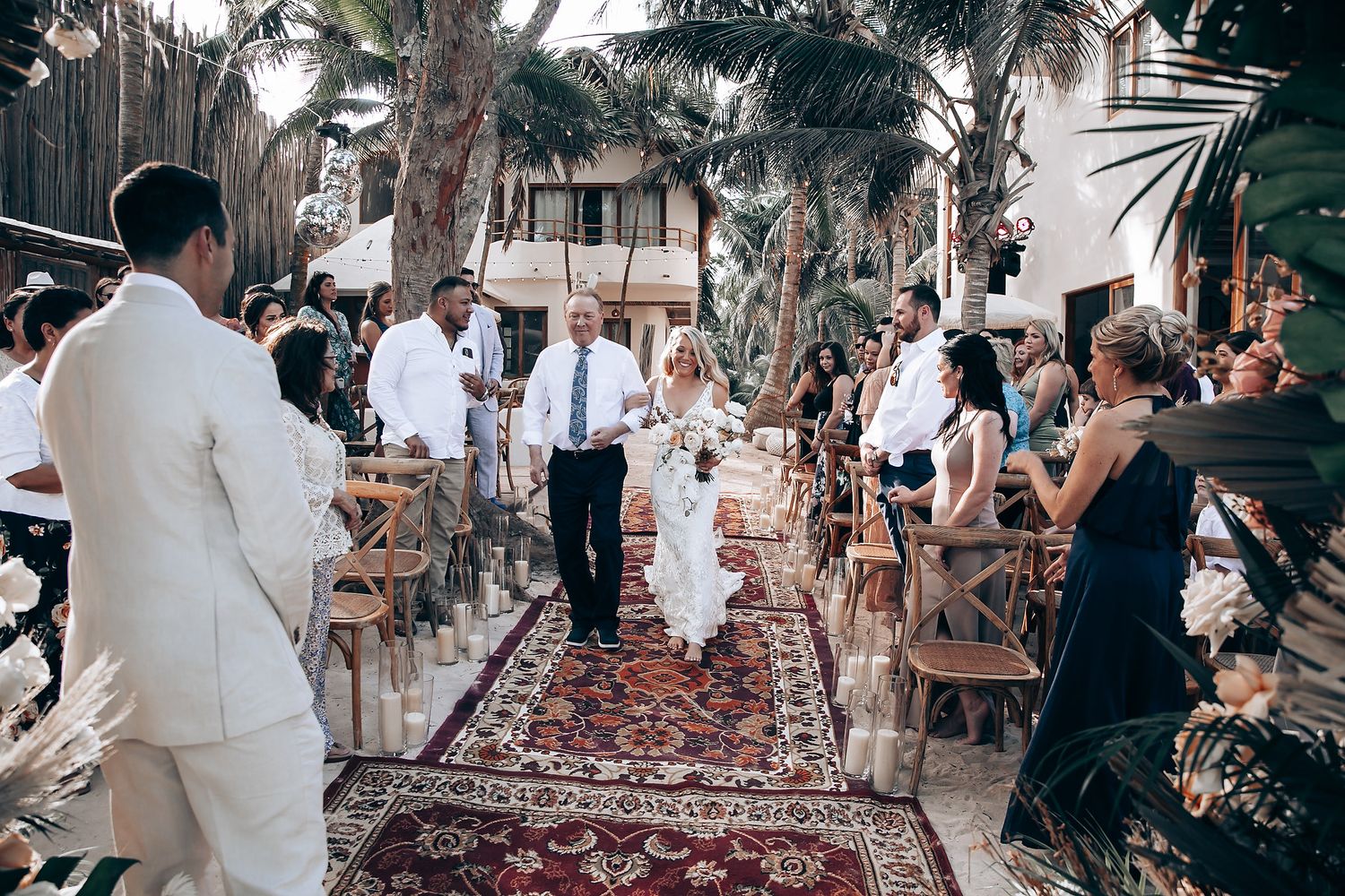 Outdoor wedding ceremony with Persian rug aisle runner and floral arch surrounded by palm trees.