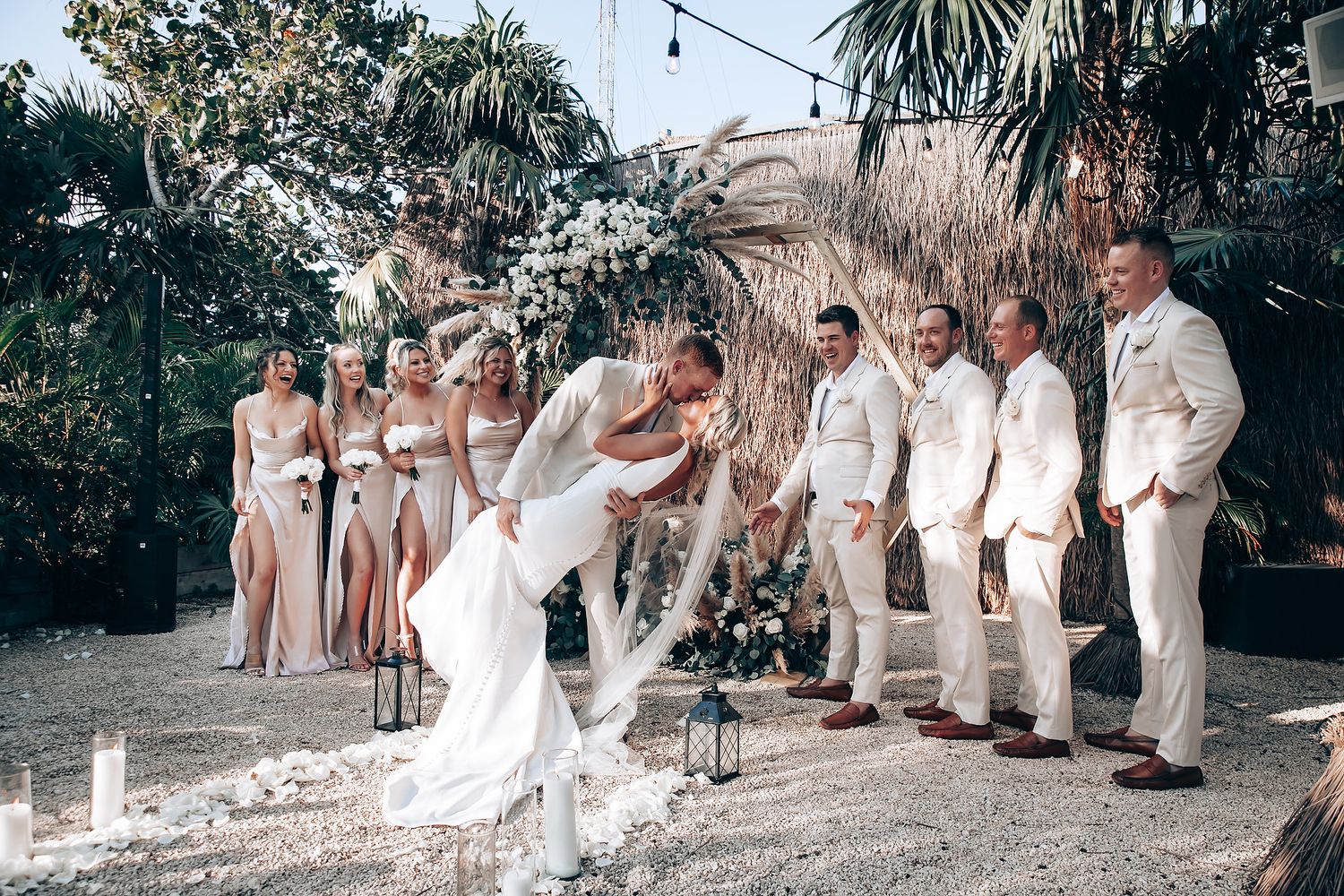 Garden wedding ceremony with guests seated on wooden chairs under palm trees.