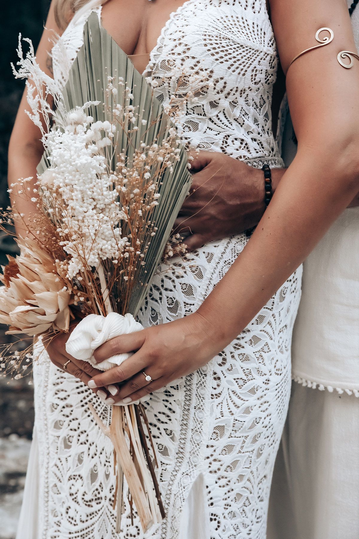 Close-up detail of delicate white lace wedding dress with dried floral bouquet held against it.
