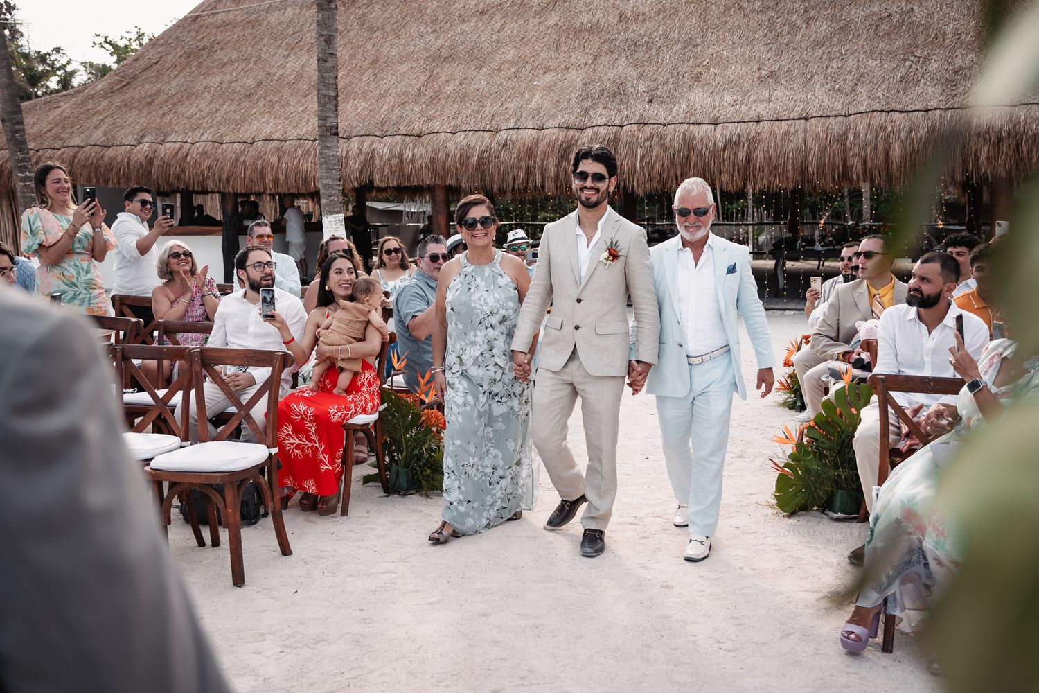 Groom walks down beach wedding aisle with parents in tropical outdoor ceremony.